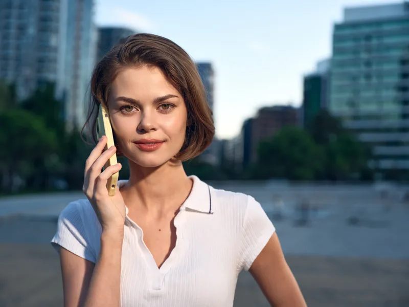 A cheerful professional woman stands in an outdoor urban setting, talking on a smartphone. She smiles warmly, projecting a confident, approachable vibe that suggests productive, modern business