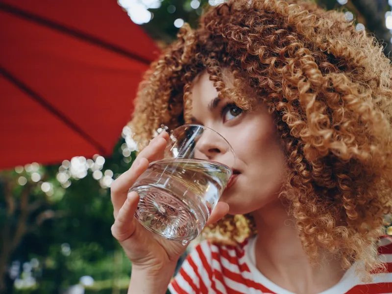Close portrait of a young woman with curly hair drinking water from a glass, outdoors in bright light, wearing a striped shirt and a calm, focused expression.