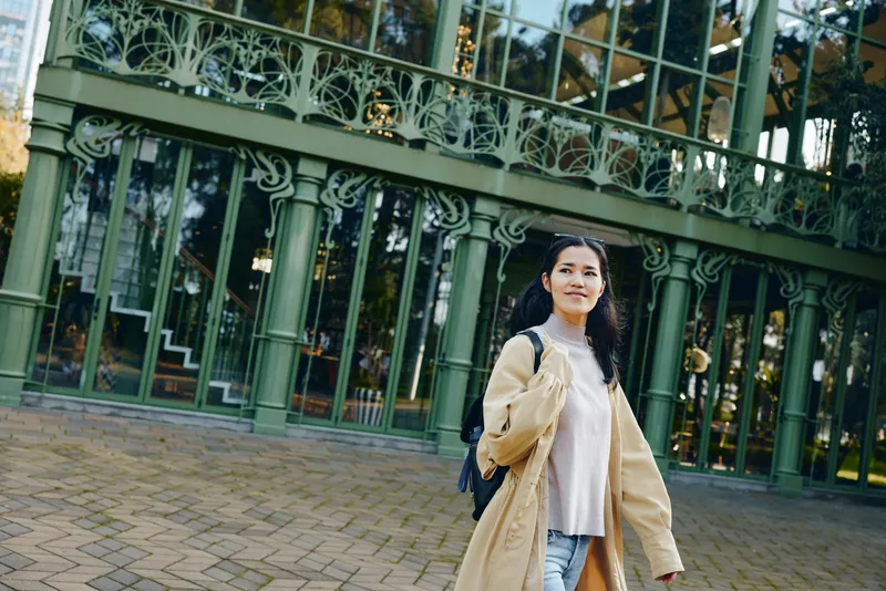 Woman in a beige coat and white sweater stands on a brick pavement in front of a glass building, showcasing casual street fashion, outdoor style, and a friendly smiling mood