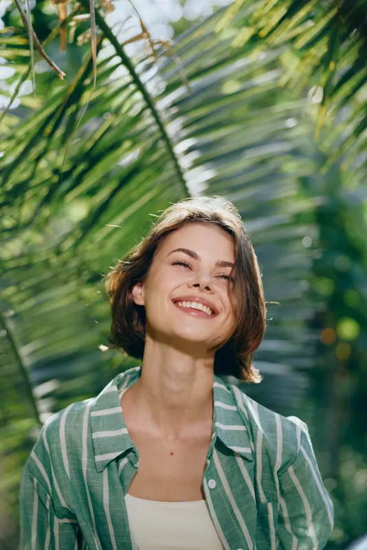 Smiling woman wearing a green striped jacket stands among lush tropical plants under bright sunlight, creating a fresh, summery portrait.