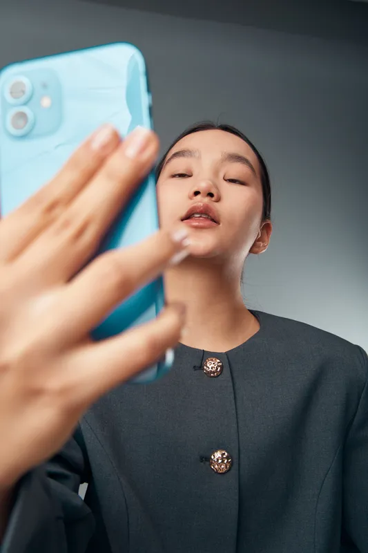 Young woman taking a selfie with a blue smartphone, wearing a dark buttoned jacket, looking confident against a plain gray background for social media or personal use.