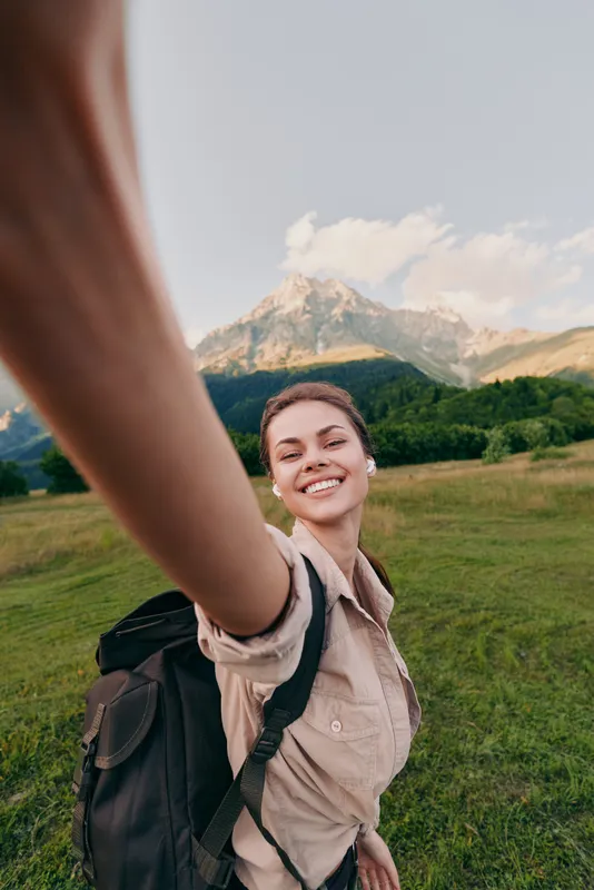 Woman outdoors with backpack smiles for a selfie during a hike in a grassy valley beneath mountains, enjoying nature and travel in bright daylight, casual lifestyle