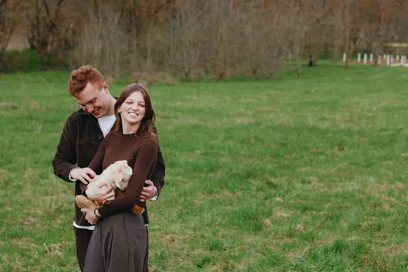 A couple outdoors in a grassy field cuddle a friendly goat kid, smiling at the camera. A natural, candid lifestyle moment illustrating affection, happiness, and companionship with animals in a rural