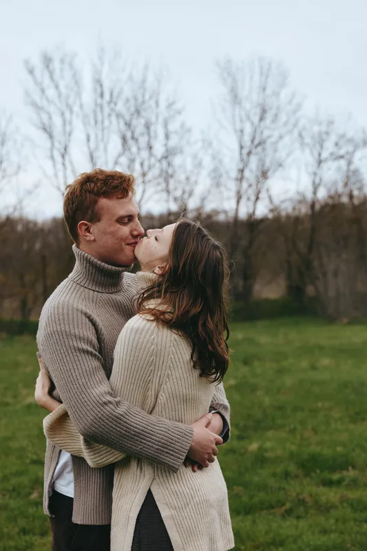 Couple kissing outdoors in cozy sweaters, romance and embrace in a field of nature, love shines through a warm, seasonal moment with soft light and gentle breeze in a rural setting.