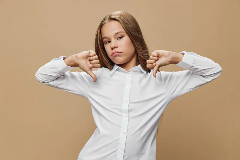 Girl in white shirt showing dislike gesture with both hands thumbs down standing on beige background. Young female expressing negative emotion with confident pose and serious face.