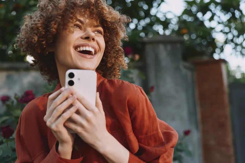 Joyful woman outdoors holds a phone, smiling with curly hair in a red sweater. Bright natural light and greenery frame a candid lifestyle moment focused on happiness and everyday joy
