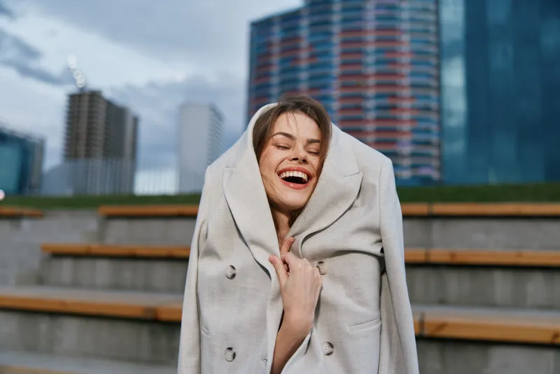 Young woman wrapped in beige coat smiling outdoors in urban setting with modern buildings and overcast sky