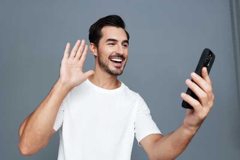 Cheerful smiling man waving hand while using mobile phone for video call, standing against a plain gray background. Modern lifestyle concept.