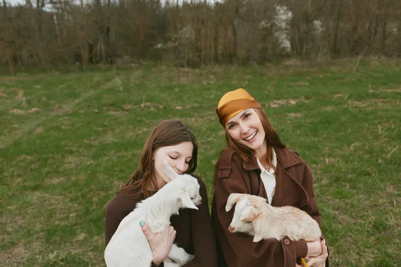 Two women in brown coats cradle playful goat kids in a grassy field, smiling as gentle interaction unfolds. A lifestyle moment outdoors showing companionship and caretaking in a rustic setting.