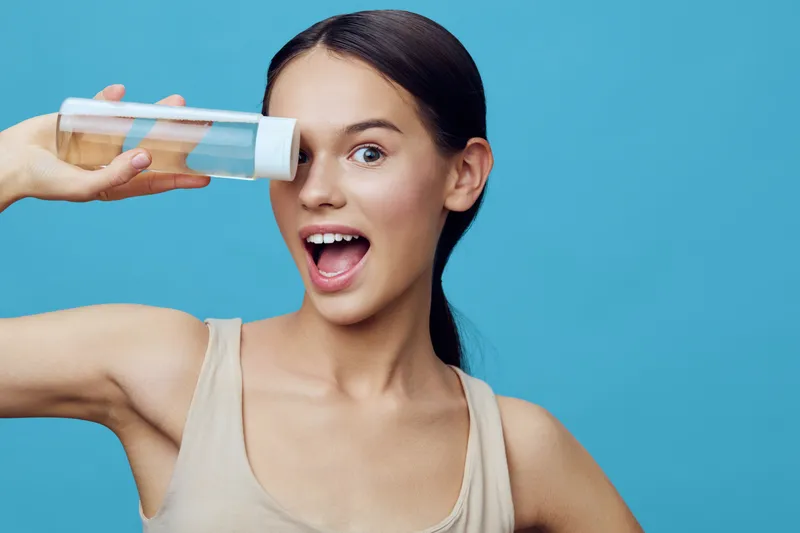 Young woman with long hair playfully holding a clear bottle against a blue background, showcasing joy and vitality while promoting healthy lifestyle choices