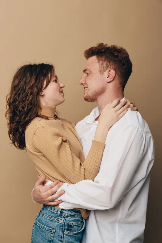 Young couple embraces and looks into each other’s eyes with smiles. Woman in beige sweater and man in white shirt share intimate moment on neutral background. Concept of love and connection.