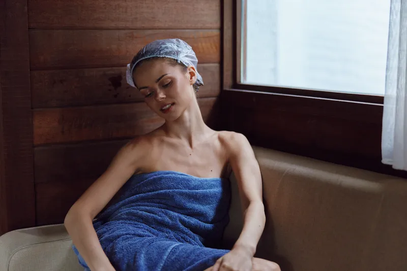 young woman wrapped in blue towel relaxes inside wooden sauna with shower cap on head, enjoying peaceful and calm atmosphere, natural light from window