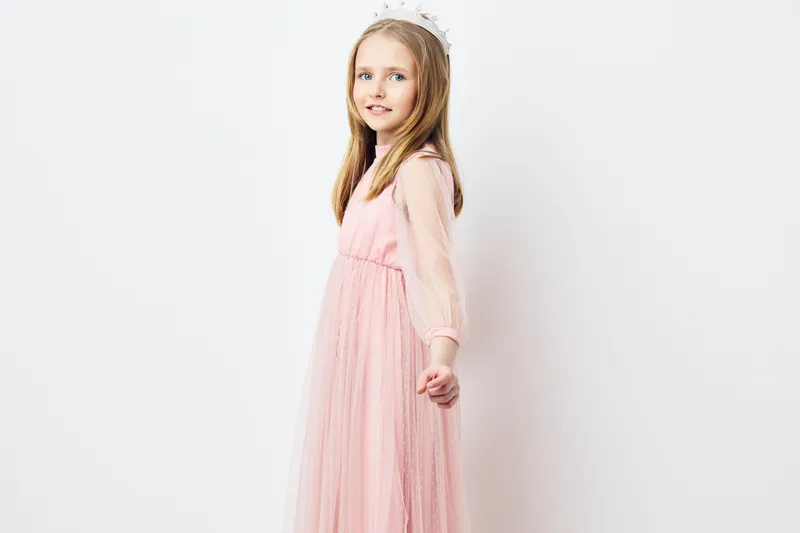 Cute girl in a pink dress with a tiara, smiling gently, stands against a simple white background, embodying joy and innocence in a vibrant, playful setting