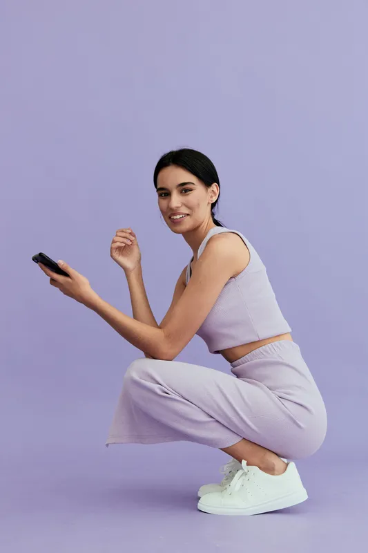young woman squatting in casual purple outfit with white sneakers, smiling gently against light lavender background, showing relaxed and cheerful mood