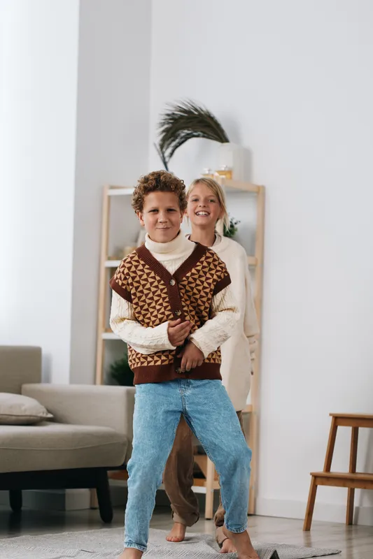 Authenticity and playful school lifestyle are shown as two children indoors, one wearing a knitwear vest and jeans, the other smiling in natural light.
