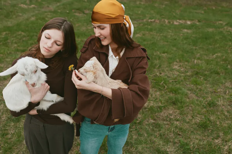 Two young women hold baby goats outdoors on a grassy field while smiling and enjoying a relaxed lifestyle moment in casual autumn clothing.