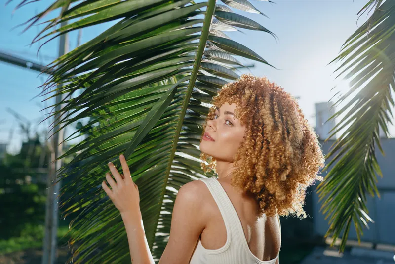 smiling woman with curly hair stands near tropical palm leaves outdoors under bright sunlight, wearing white sleeveless top, enjoying warm weather and natural beauty.