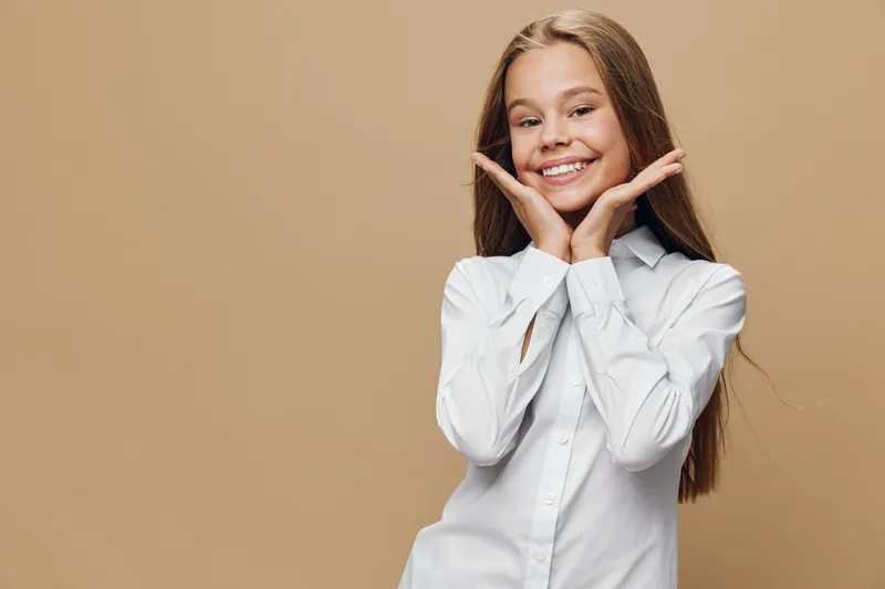 Girl with long hair smiling and posing with hands near face in white shirt isolated on beige background. Charming child, happy expression and casual style portrait.