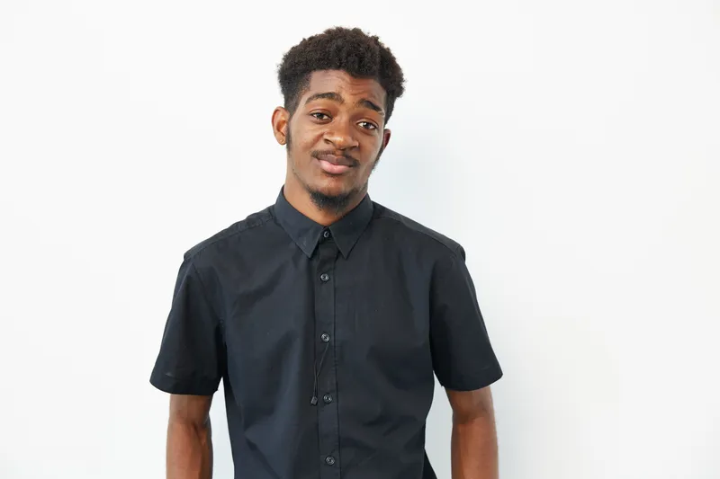 Portrait of a black man wearing a black shirt, showcasing a relaxed expression and a modern hairstyle. The background is minimalistic, enhancing his features.