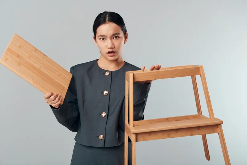 Woman holding broken wooden chair and detached plank indoors, surprise and concern on face, furniture damage repair concept with casual outfit on gray background.