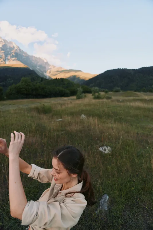 Young woman outdoors in mountain meadow, smiling with raised arms, casual outfit, natural light, peaceful landscape, lifestyle concept, fresh air, sunny day