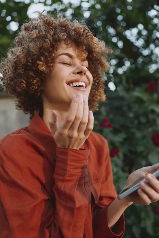 Young woman with curly hair smiles while holding a smartphone outdoors, enjoying a bright moment of connection, laughter, fresh air, and casual everyday life