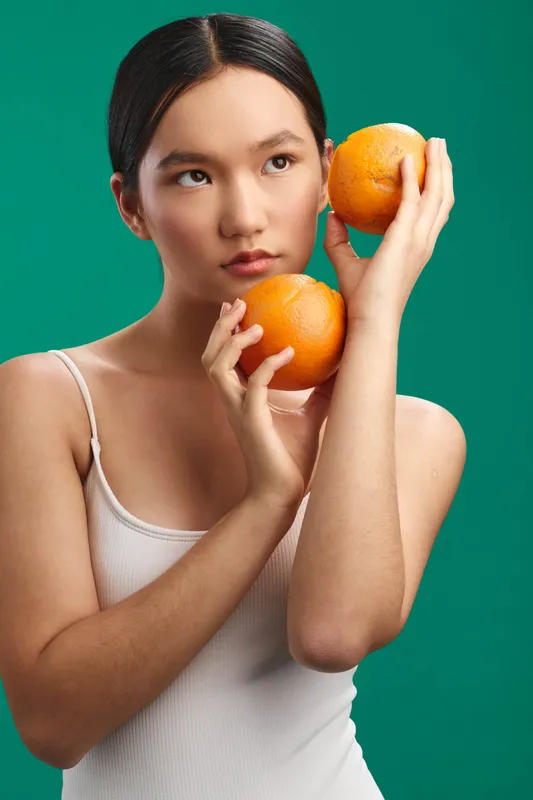 asian woman holds bright oranges against a green backdrop, presenting fresh fruit with a calm, thoughtful expression. clean styling and soft lighting create a healthy, vibrant lifestyle mood.