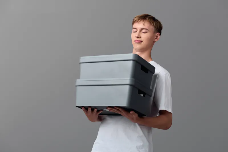 Delivery scene featuring a man in a white shirt holding a stack of grey boxes, ready for shipment, emphasizing packaging, logistics, and courier service in a clean studio setting.