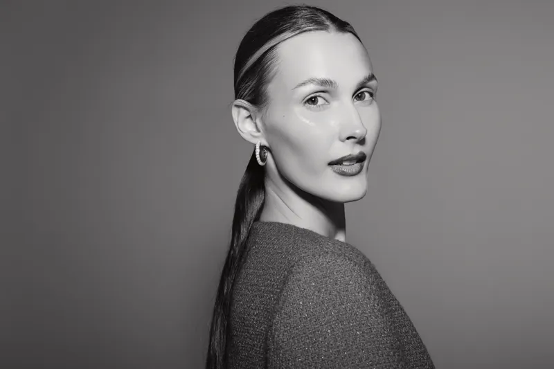Portrait of a fair skinned Caucasian woman with slicked back hair wearing earrings and a textured top in a black and white studio setting.