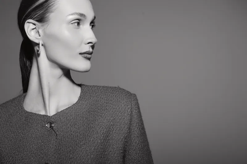 Elegant Caucasian woman with pale skin in textured jacket looks sideways against grey background. Portrait in black and white studio lighting with minimalistic style.