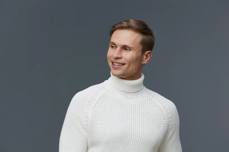 Smiling young man with short brown hair wearing white knitted turtleneck sweater looking to the side portrait isolated on grey background. People lifestyle concept