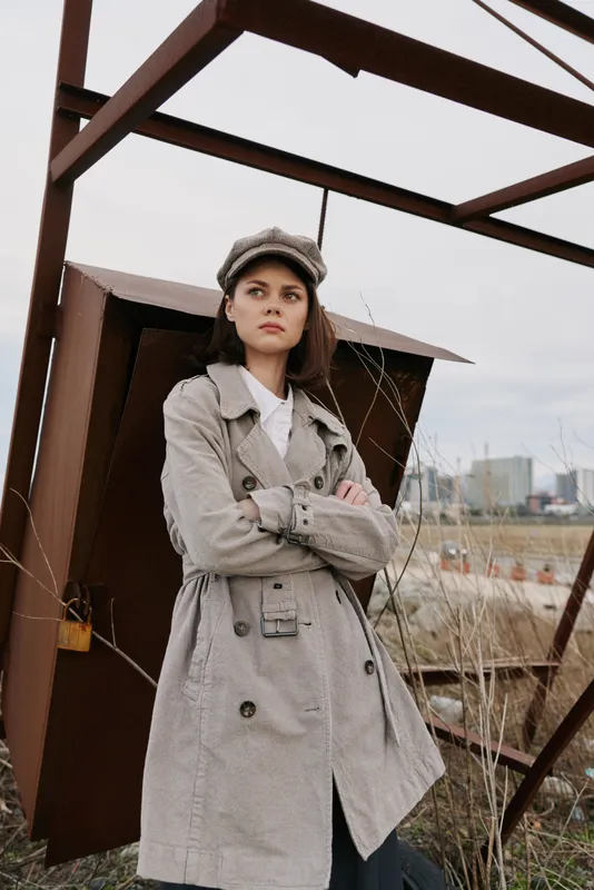 Young woman in checkered trench coat and hat stands with crossed arms near industrial structure outdoors. Overcast sky and urban background create moody atmosphere in fashion portrait.