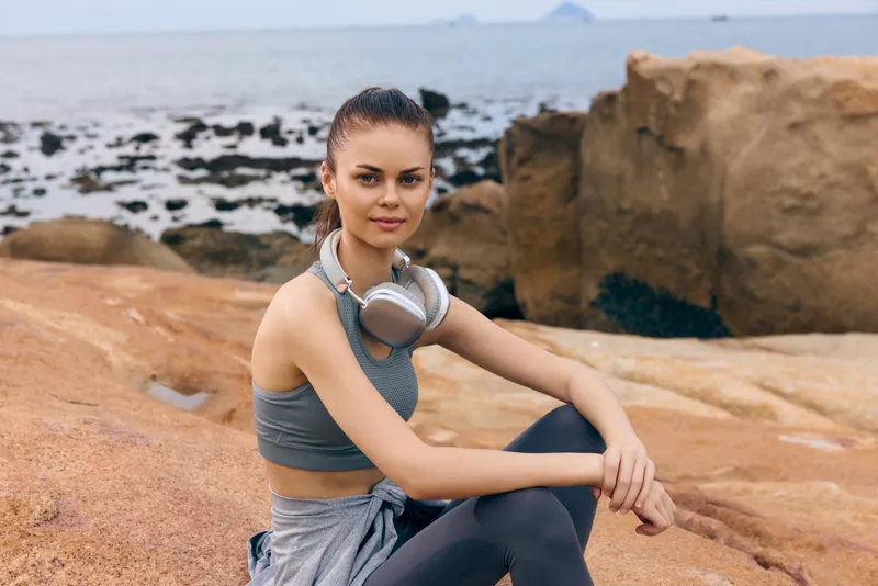 Young woman in active wear with headphones, sitting on rocky beach, enjoying nature and music, showcasing fitness lifestyle and relaxation
