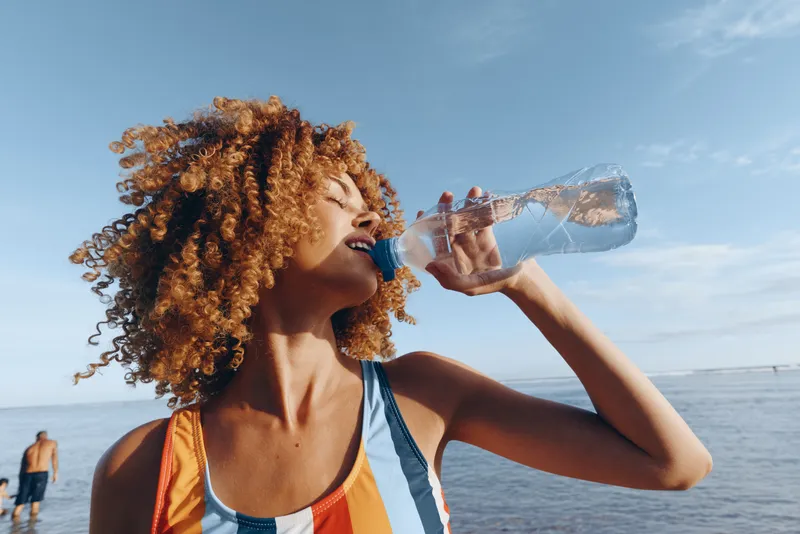 Woman with curly hair drinking water on the beach, ocean horizon behind, summer outdoor hydration, sunlight on skin, close side view, energetic wellness