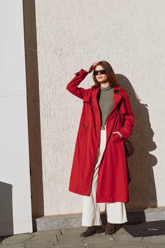 A stylish woman wearing a red trench coat stands against a sunlit white wall, paired with a green top and white wide leg pants, exuding confident, casual elegance.