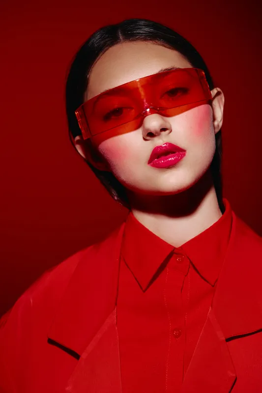 Young woman in red outfit with slick hairstyle and glossy lips wears futuristic red sunglasses. Bold look on seamless red background. Concept for beauty creative fashion futuristic.