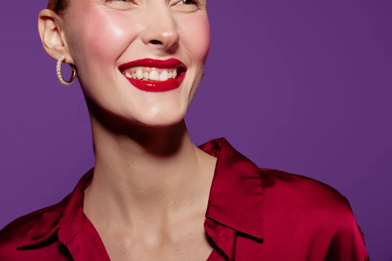 Close-up of smiling woman with red lipstick and hoop earrings wearing red satin blouse on purple background. Concept of beauty, fashion, makeup, and happiness.
