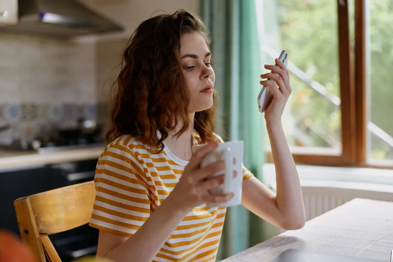 Casual woman indoors, focused on smartphone, natural window light, relaxed lifestyle moment, striped shirt, home atmosphere, candid portrait of everyday technology use