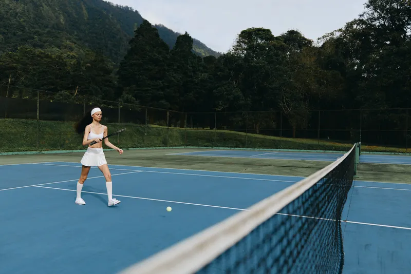 Young female tennis player in a white sports outfit joyfully playing on a blue outdoor court surrounded by lush greenery in a serene environment