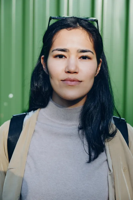 Woman with a neutral expression outdoors against a green corrugated backdrop, wearing a light turtleneck and beige jacket, sunglasses perched, casual lifestyle shot