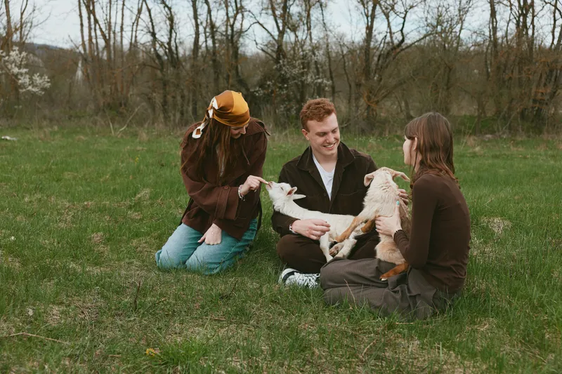 Three young adults enjoy a relaxed moment outdoors in spring while holding and playing with two baby goats on a grassy field surrounded by bare trees.