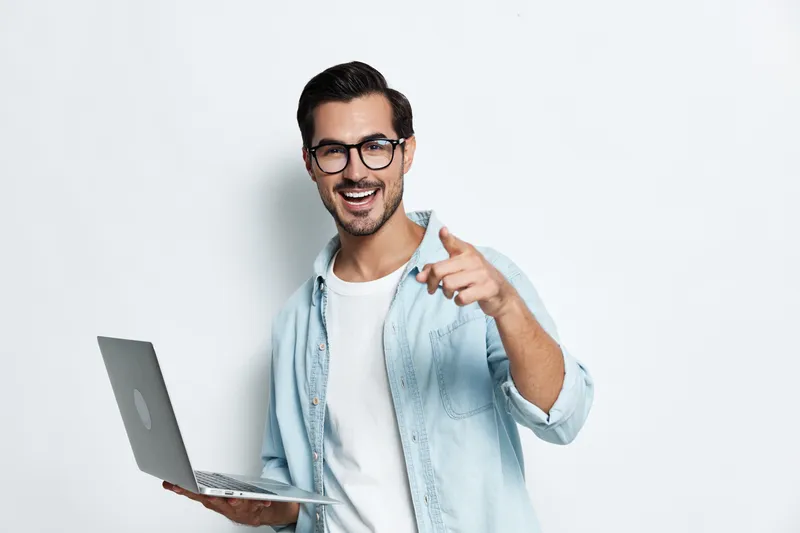 Handsome smiling young man wearing glasses and a light blue shirt, holding a laptop and pointing at the camera with a joyful expression on his face. Studio portrait.