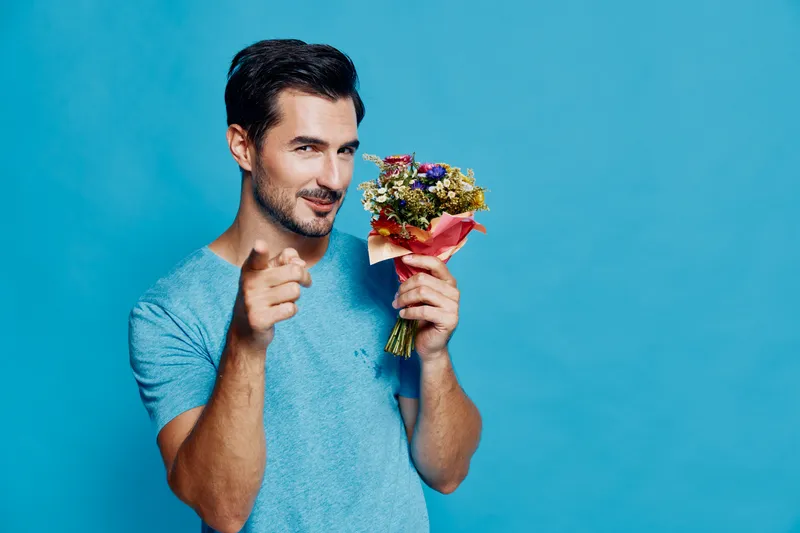 Smiling man holding a bouquet of colorful flowers while pointing at the camera, showcasing a playful and charming personality against a bright blue background.