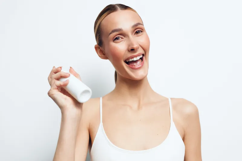 A happy young woman in a white tank top holds a white bottle while smiling brightly against a plain white background. Concept for beauty, skincare, and health products.
