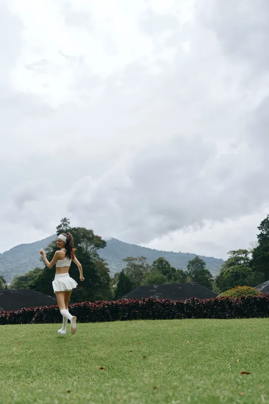 Young woman jogging in a green field, wearing a sporty outfit, with a joyful expression against a natural backdrop of mountains and clouds
