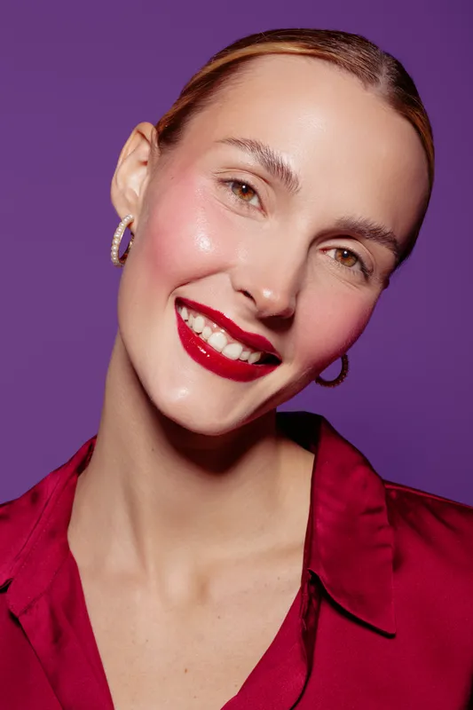 Smiling woman with red lipstick and hoop earrings poses against purple background. She wears a red blouse and has glowing skin, conveying confidence and beauty in a studio portrait.