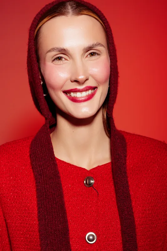 Smiling woman with red lips and shiny skin wears red sweater and hood against red background. Portrait of happy female model with natural makeup and bright festive mood.