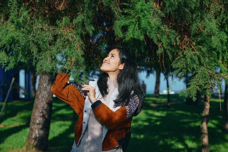 Woman enjoying nature outdoors beneath a leafy umbrella, reaching toward hanging branches with a joyful smile, showcasing casual fashion and spontaneous moment in a sunlit park