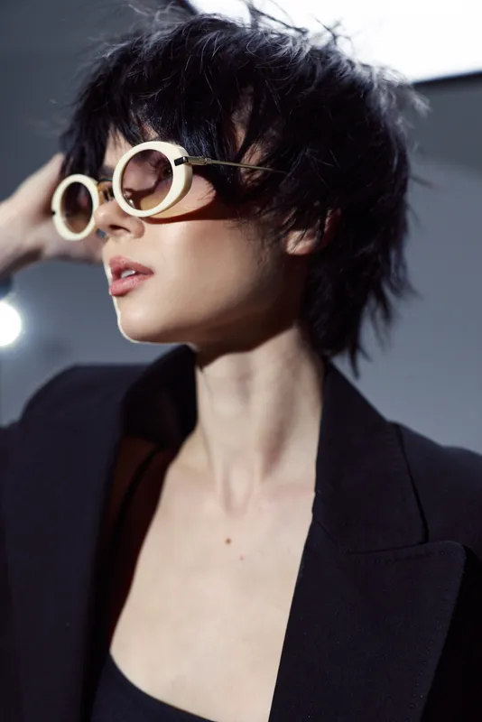Stylish woman wearing round sunglasses and a black jacket with short dark hair and a confident expression.