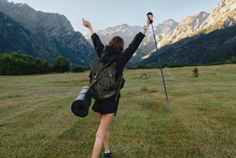 A cheerful hiker with a backpack stands in a wide meadow beneath towering mountains. She raises a trekking pole, smiling as she embraces a moment of outdoor exploration and freedom.